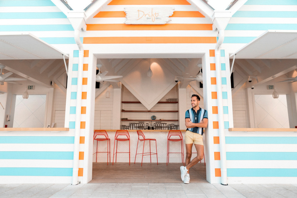 A man in a collared shirt and shorts stands in the doorway of a brightly colored outdoor bar featuring orange and light blue stripes.