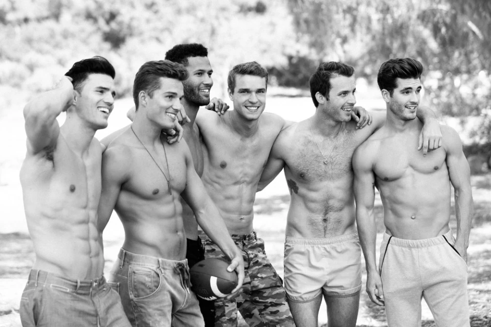 Black and white shot of six athletic adult men standing outdoors smiling with a football, wearing jeans, shorts and camouflage pants.