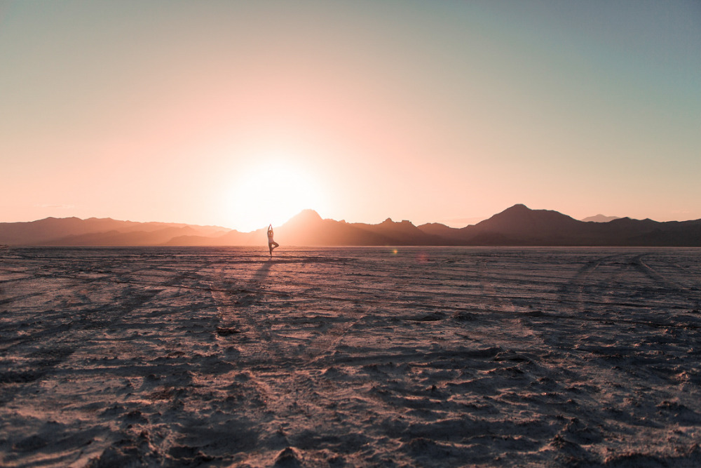 Woman in tree pose, standing on cracked earth at sunset with mountain range in background; fitness, yoga, exercise, outdoor landscape.