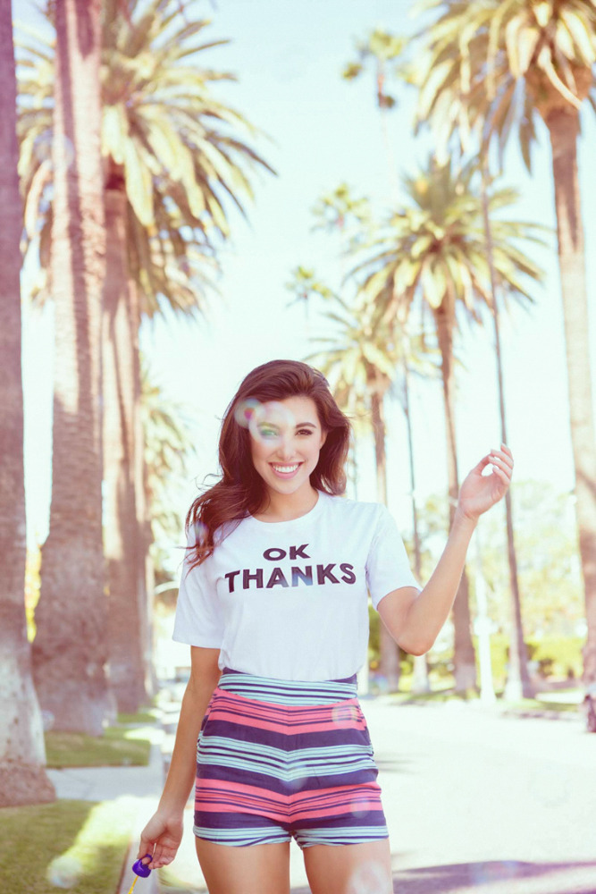 A smiling woman in a T-shirt and striped shorts stands on a palm-tree-lined street in full sunlight holding a bubble wand.