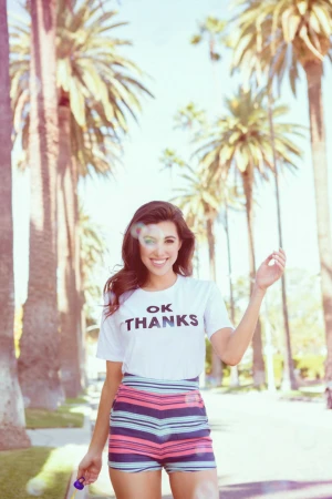 A smiling woman in a T-shirt and striped shorts stands on a palm-tree-lined street in full sunlight holding a bubble wand.