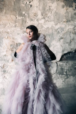 Woman in a dramatic tulle gown posing in front of a distressed wall. The dress features layers of ruffles and a high ruffled collar.