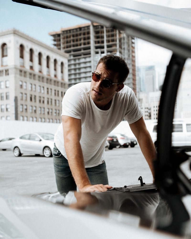 Man in white t-shirt and sunglasses leans on silver car with open hood in urban setting with city buildings in the background.