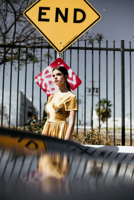 A stylish young woman poses outdoors in front of street signs and a black metal fence, wearing a gold dress in natural light.