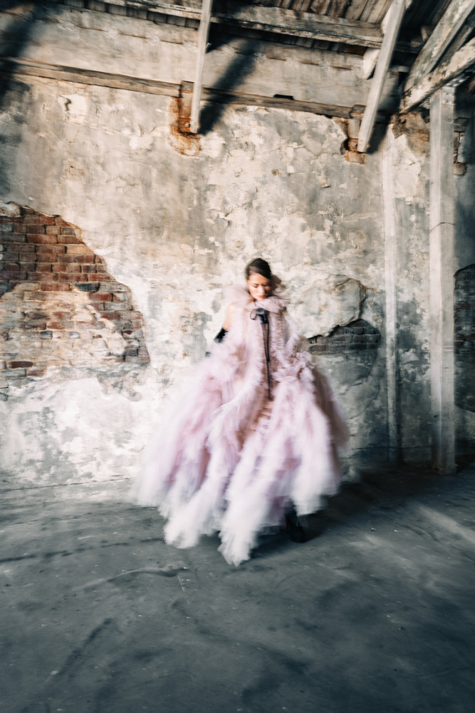 A woman poses in a large, pale pink, ruffled gown in a derelict, sparsely lit room, showcasing editorial fashion in a crumbling, old setting.