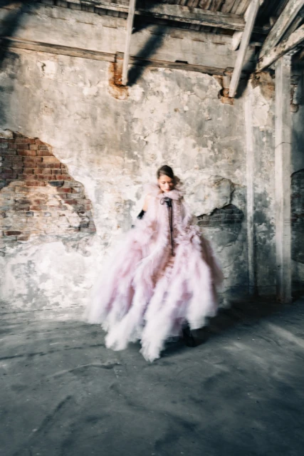 A woman poses in a large, pale pink, ruffled gown in a derelict, sparsely lit room, showcasing editorial fashion in a crumbling, old setting.