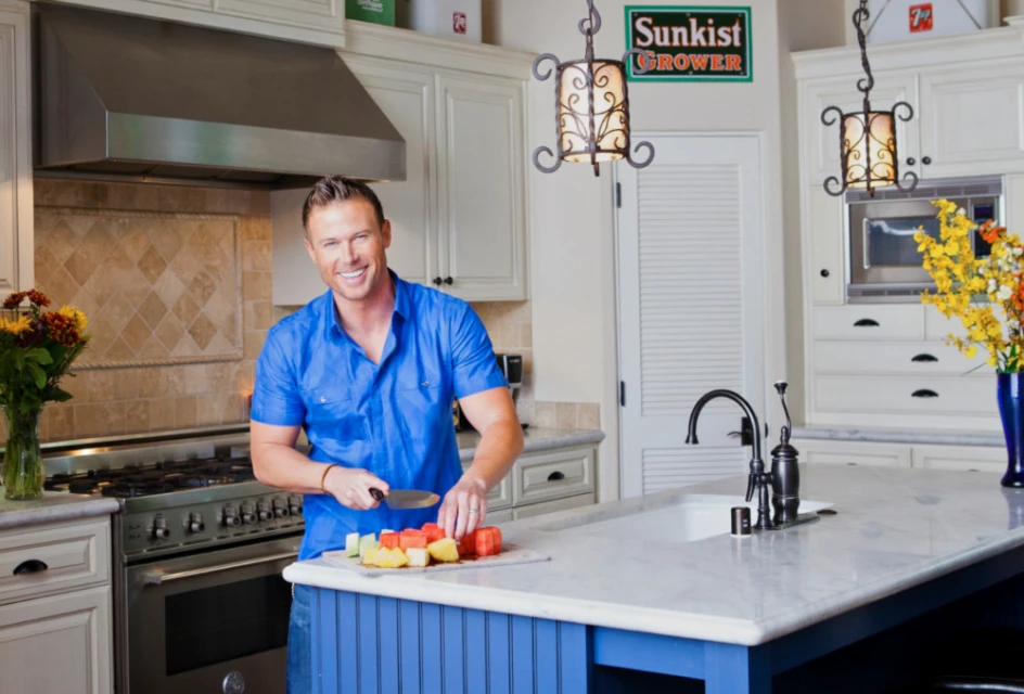 Smiling man in blue shirt cuts fruit on a kitchen island with stainless steel appliances, light wood cabinets, and a Sunkist Grower sign.