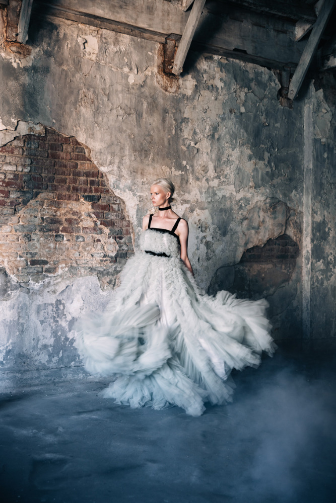 Woman in a white ruffled gown poses in a derelict indoor space with peeling plaster and atmospheric fog, fashion editorial photography.