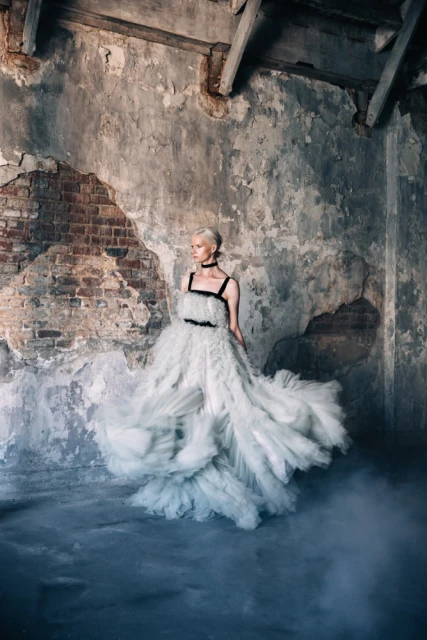 Woman in a white ruffled gown poses in a derelict indoor space with peeling plaster and atmospheric fog, fashion editorial photography.