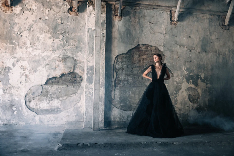 Elegant woman in a black gown poses in a dilapidated interior with cracked walls and smoke effects, showcasing high fashion.