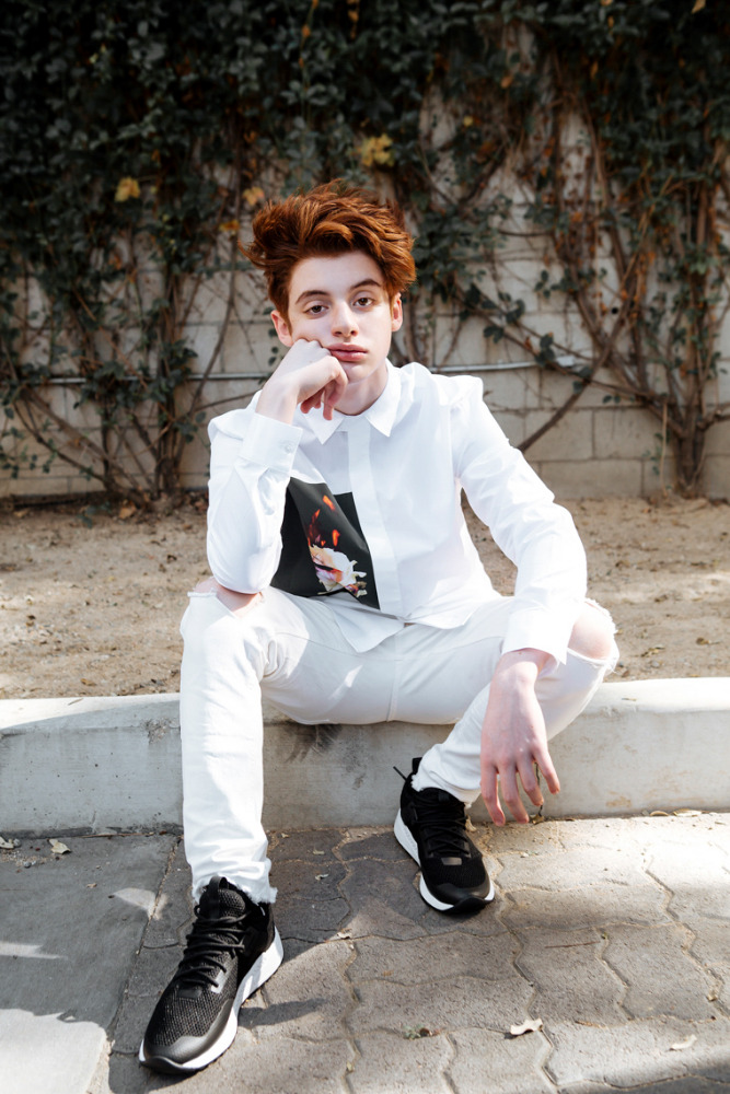 Young adult with red hair in an outdoor portrait, wearing a white collared shirt, ripped white jeans, and black sneakers, seated on a low wall.