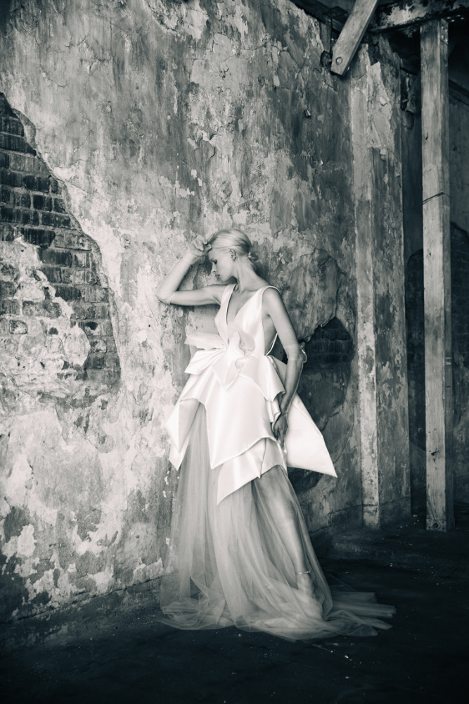 Monochrome shot of a woman in a white tiered gown posing against a textured wall in an indoor space with natural lighting.