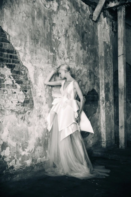 Monochrome shot of a woman in a white tiered gown posing against a textured wall in an indoor space with natural lighting.