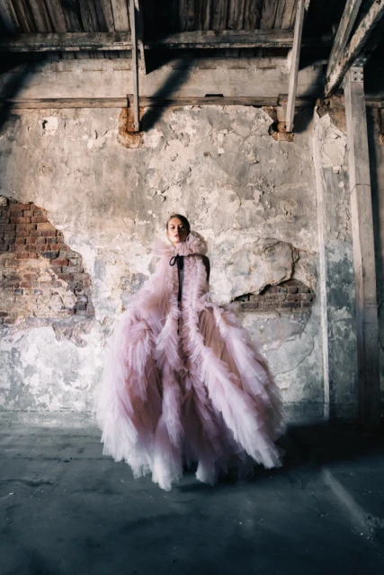 Woman in large pink gown poses in abandoned building with peeling paint and brick walls, showcasing fashion and unique setting.