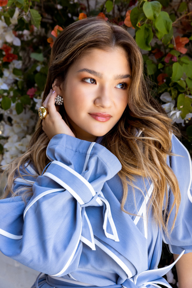 Portrait of a smiling young woman with long brown hair in a blue wrap dress, posing outdoors with flowers in the background.