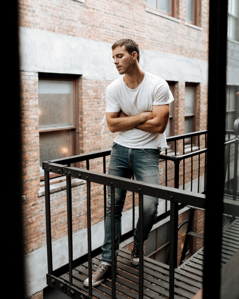 Adult man poses on a metal fire escape with a brick building facade in the background, looking down with arms crossed.