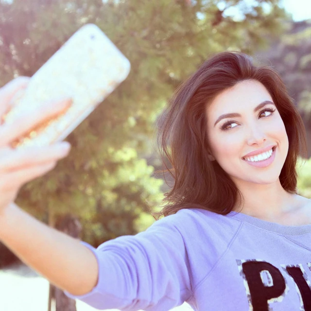 Brunette woman in purple sweatshirt smiles and takes a selfie with a smartphone outside against a natural green background.