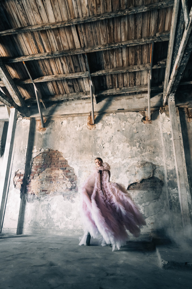 Woman in ruffled pink gown stands in derelict room with peeling walls and wooden rafter ceiling, creating a dramatic fashion portrait.
