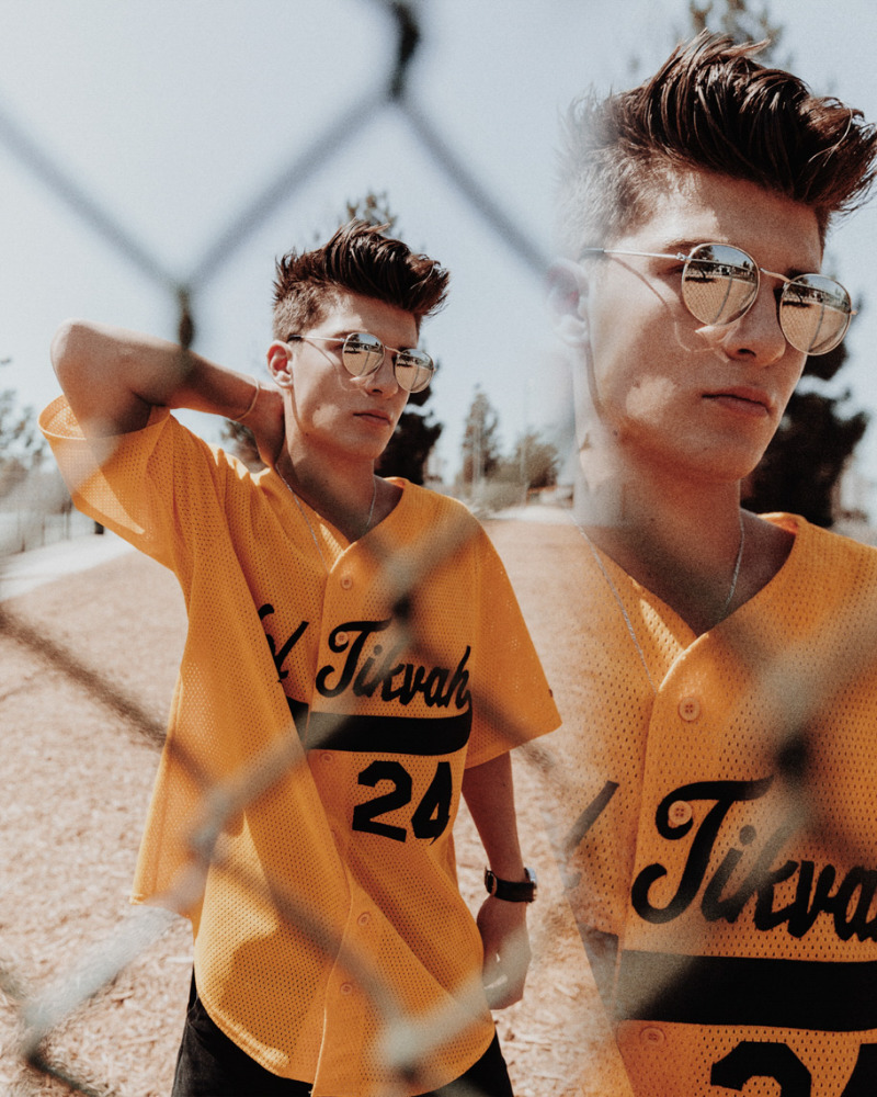 Portrait of a young adult male in a yellow baseball jersey with mirrored sunglasses, posing outdoors with a chain link fence in the foreground.