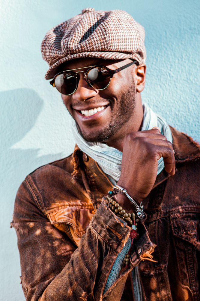 Smiling African American man in tweed cap and sunglasses posing against a blue wall, wearing a distressed brown denim jacket.