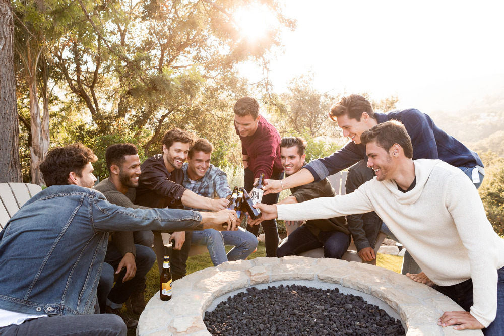 Group of eight young adult men toasting bottled beers in a grassy backyard by a stone fire pit, trees and sunlight in background.