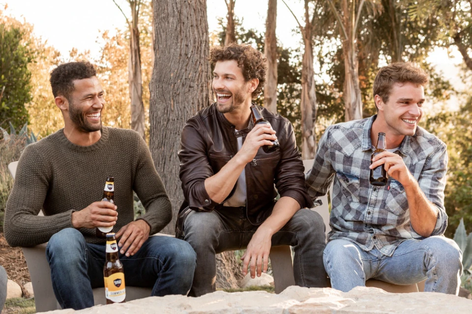 Three adult males sitting outdoors, holding beer bottles, smiling in natural light. Casual wear, friendship, leisure, relaxation.