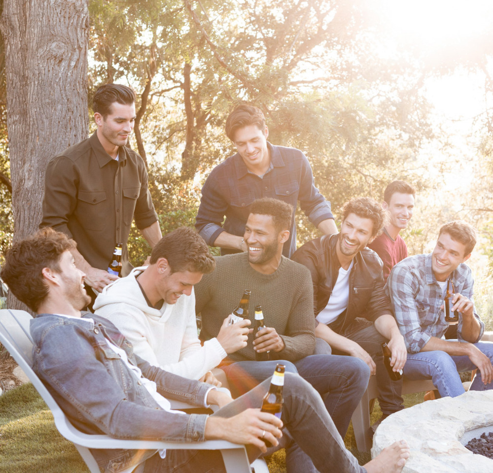 Group of eight men outdoors in casual clothing, holding bottles, sitting around a fire pit on a sunny day with trees in background.