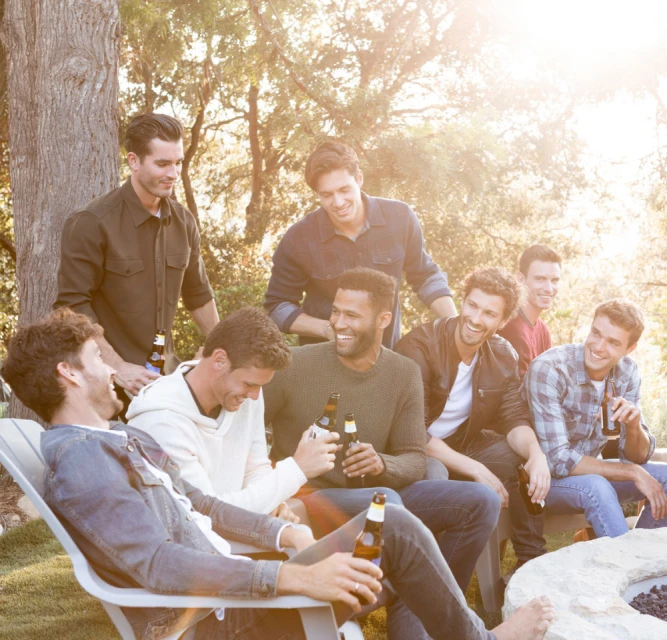 Group of eight men outdoors in casual clothing, holding bottles, sitting around a fire pit on a sunny day with trees in background.