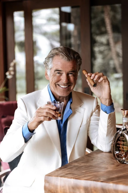Smiling adult man in a white suit jacket holds a drink and cigar inside, with greenery visible through windows in the background.