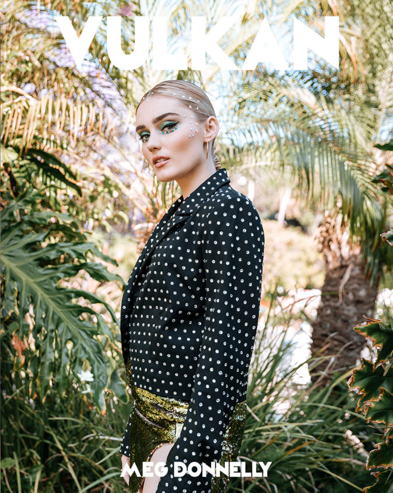 Meg Donnelly poses against a backdrop of vibrant green tropical foliage, wearing a black and white polka dot jacket and gold sequined skirt.