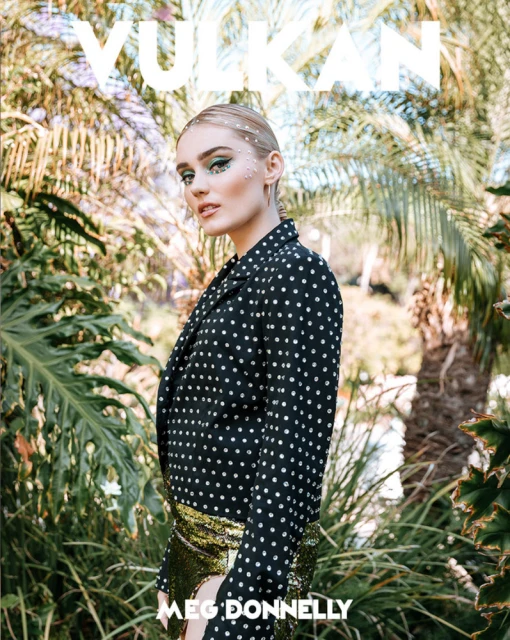 Meg Donnelly poses against a backdrop of vibrant green tropical foliage, wearing a black and white polka dot jacket and gold sequined skirt.