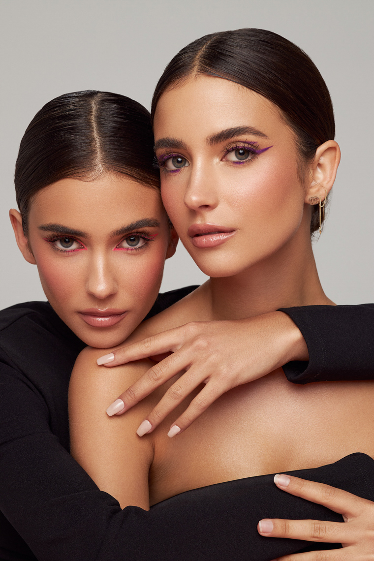 Close-up studio shot features two adult females with dark hair, dramatic makeup, and dark-sleeved tops against a neutral backdrop.