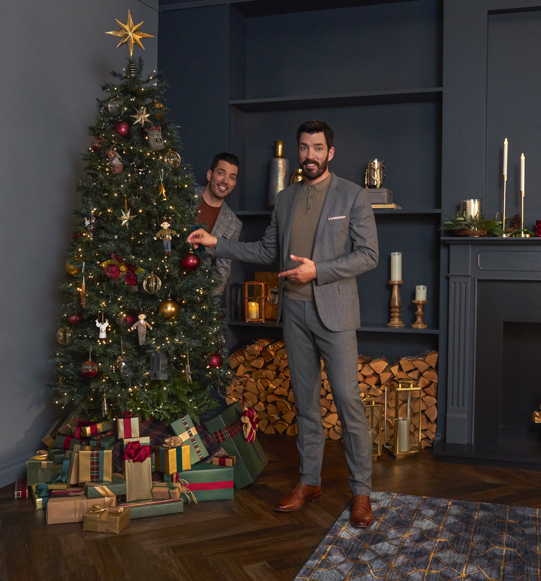 Two adult men in a holiday setting; one is decorating a Christmas tree, the other is peeking from behind it. Gifts are under the tree.