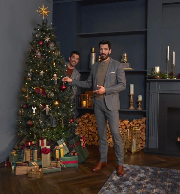 Two adult men in a holiday setting; one is decorating a Christmas tree, the other is peeking from behind it. Gifts are under the tree.