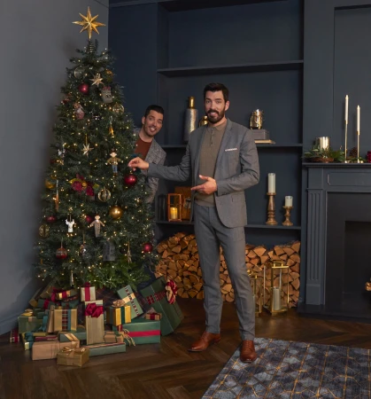 Two adult men in a holiday setting; one is decorating a Christmas tree, the other is peeking from behind it. Gifts are under the tree.