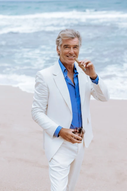 Pierce Brosnan poses on a beach wearing a white suit and blue shirt, holding a cigar and glass; ocean waves in background.