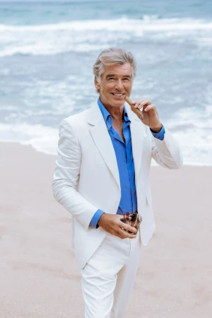 Pierce Brosnan poses on a beach wearing a white suit and blue shirt, holding a cigar and glass; ocean waves in background.
