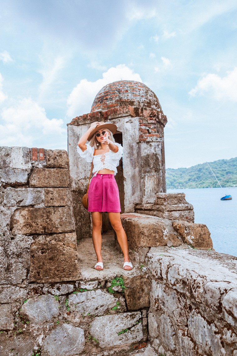 Woman in a pink skirt and white top poses on a stone fortress overlooking the ocean, wearing a hat and sunglasses on a sunny day.