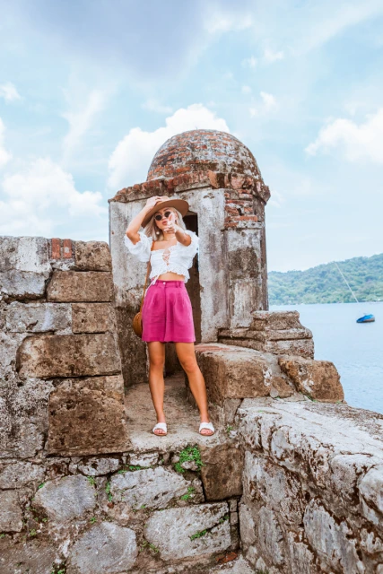 Woman in a pink skirt and white top poses on a stone fortress overlooking the ocean, wearing a hat and sunglasses on a sunny day.