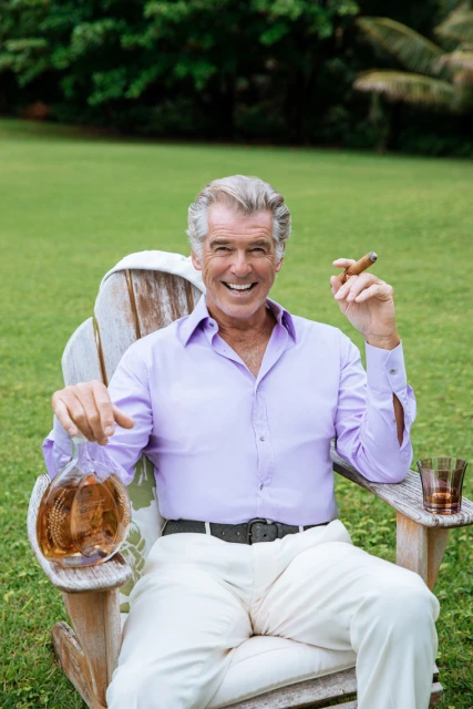 Attractive silver-haired man in lavender shirt smiles sitting in lawn chair with decanter, glass of liquor, and cigar outdoors.