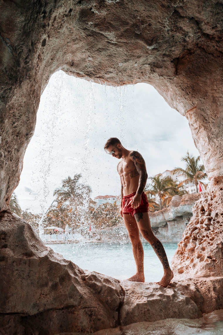 Athletic man with tattoos stands in red swim trunks near a cave opening with water and palm trees in the background on a sunny day.
