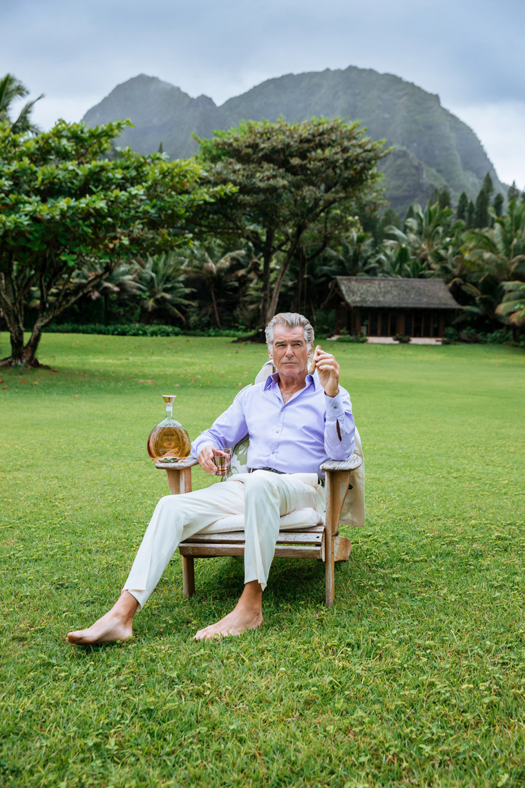 Mature man sits in a wooden chair on a green lawn with a bottle and drink in a tropical outdoor setting with mountains in the distance.