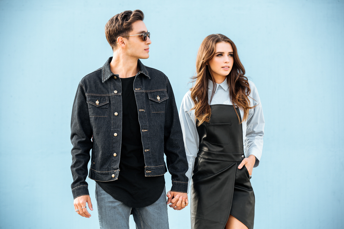 Young couple in fashionable denim and leather attire holds hands. Stylish pair poses against a light blue background, looking to the side.
