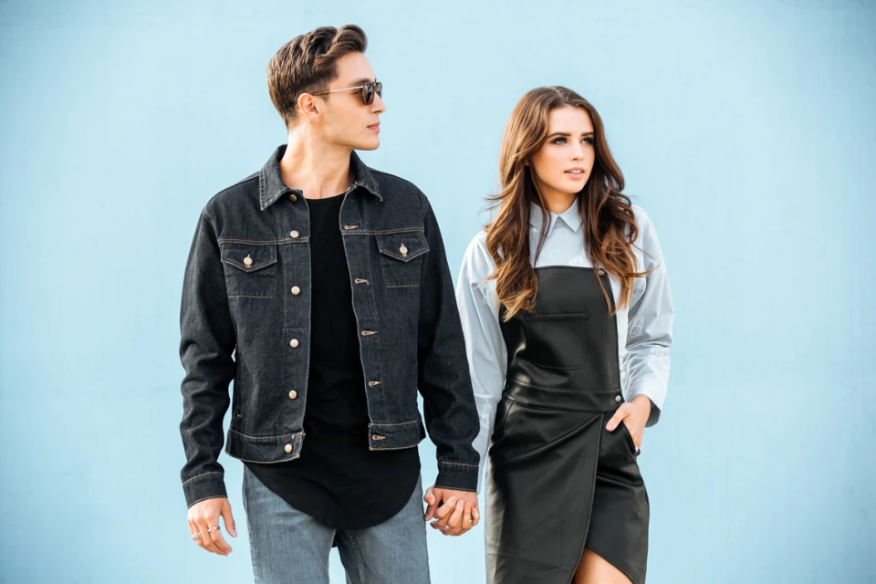 Young couple in fashionable denim and leather attire holds hands. Stylish pair poses against a light blue background, looking to the side.