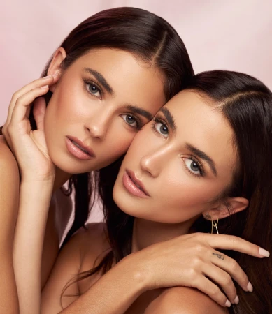 Two women with long dark hair posing closely together against a soft pink backdrop, highlighting natural makeup.