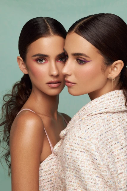 Close-up portrait of two women with dark hair and neutral makeup against a teal background, one wearing a jacket and the other a dress.