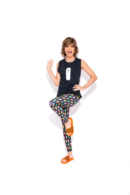 Full shot of an adult woman in a black tank top and patterned leggings, standing on one leg with an arm raised against a white backdrop.