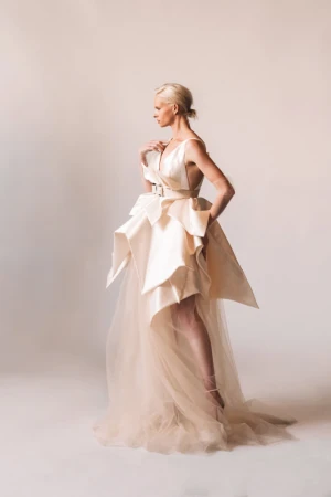 Model in an asymmetrical cream satin dress with structured peplum and tulle skirt standing against a pale seamless background.