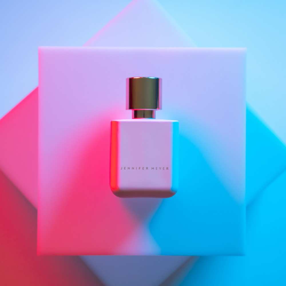 Rectangular pink perfume bottle with gold cap sitting on a geometric platform in pink and blue lighting, featuring 'Jennifer Meyer' text.