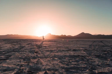 Silhouette of a person doing yoga on a vast flat salt flat during a bright sunset with distant mountains.